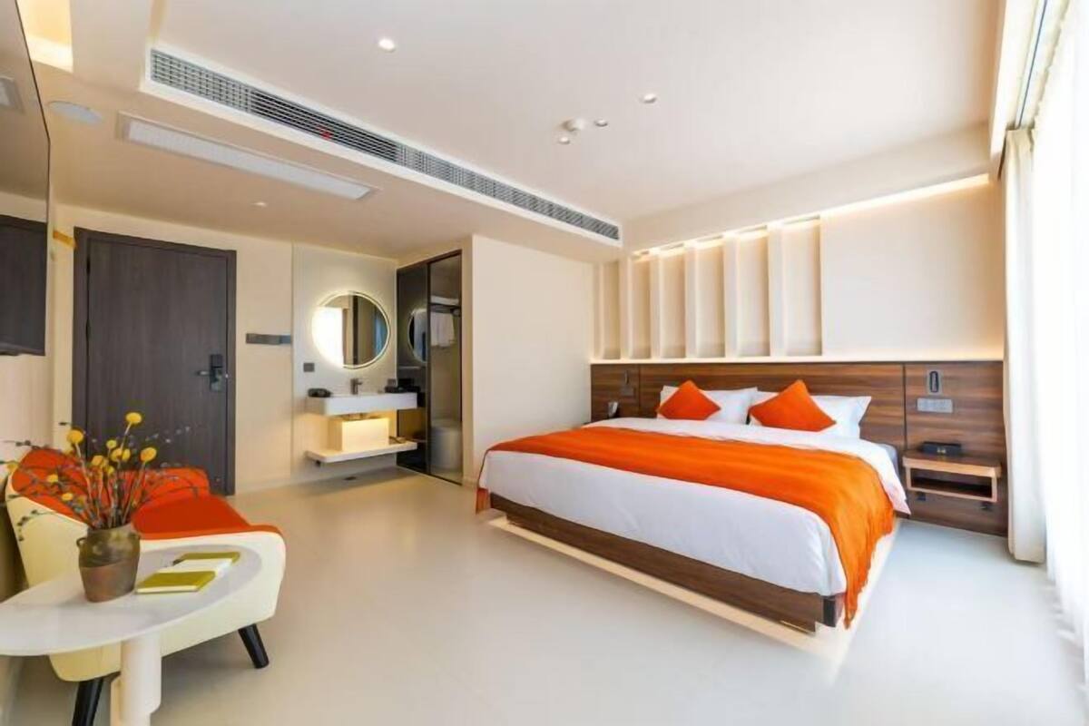 A modern hotel room with a large bed, a bedside table, a built-in wardrobe, and a small seating area with a table and chairs.