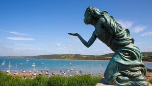 Una estatua que domina una zona costera con barcos y colinas al fondo.