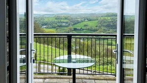Un balcón con mesa y sillas de vidrio, con vistas a campos verdes y colinas.