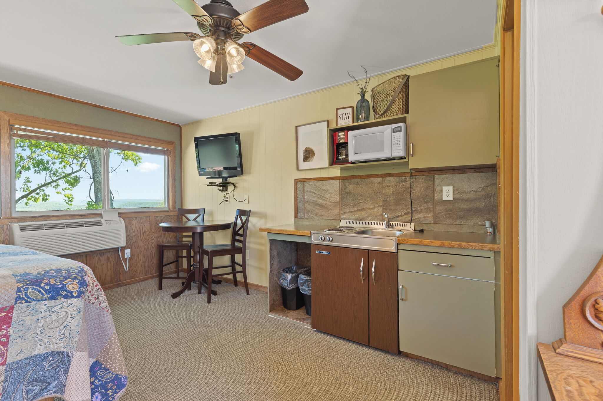 A compact room with a bed, a dining table, a kitchen area with a microwave, and a ceiling fan.