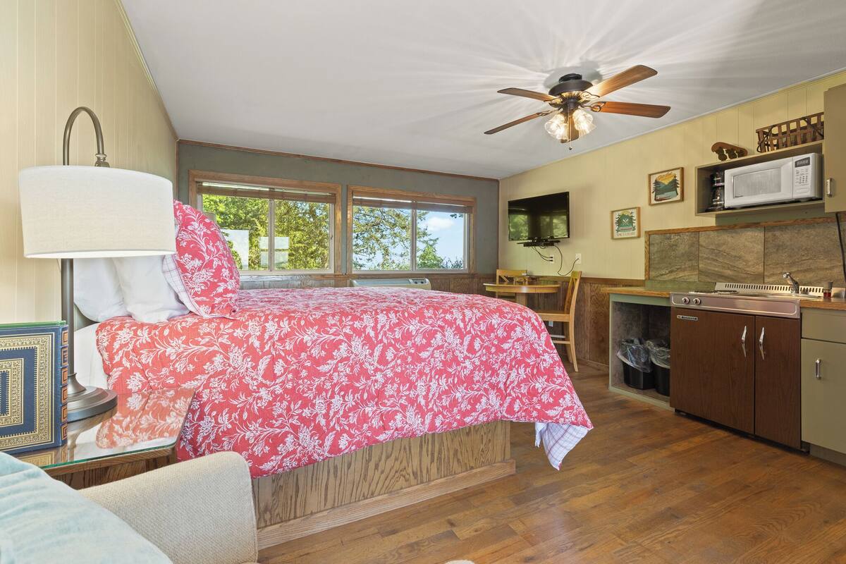 Un dormitorio con cama, escritorio, silla, ventilador de techo, ventana y una zona de cocina con microondas y armarios.