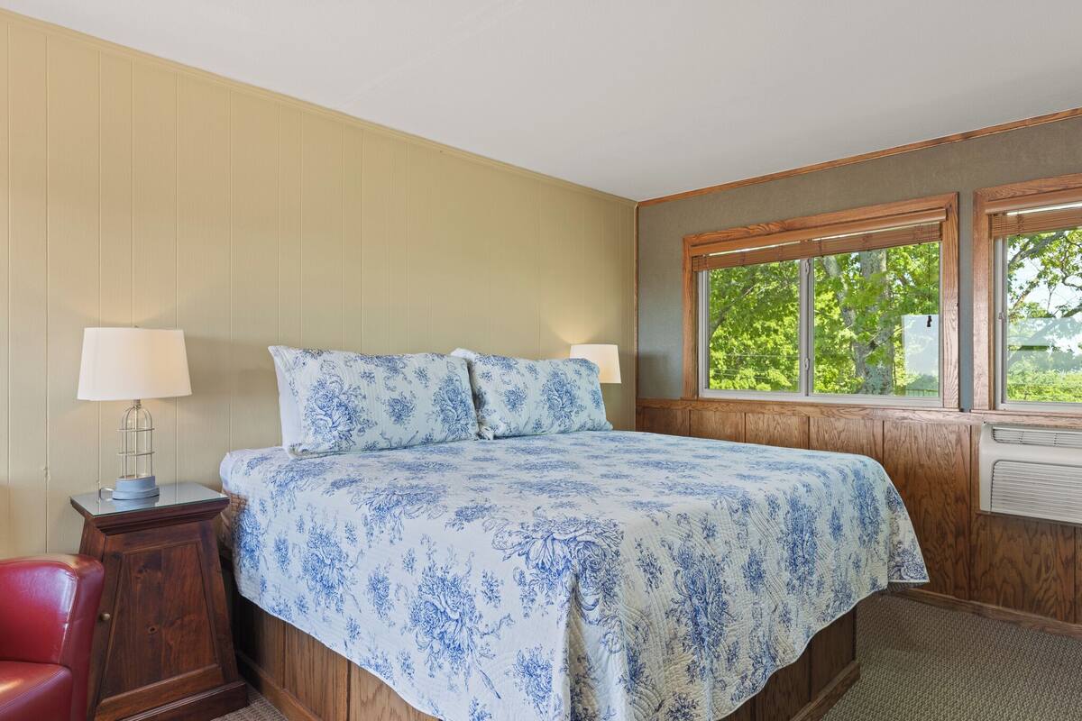 Un dormitorio con cama, dos lámparas de noche, una ventana con vistas a árboles y un cabecero de madera.
