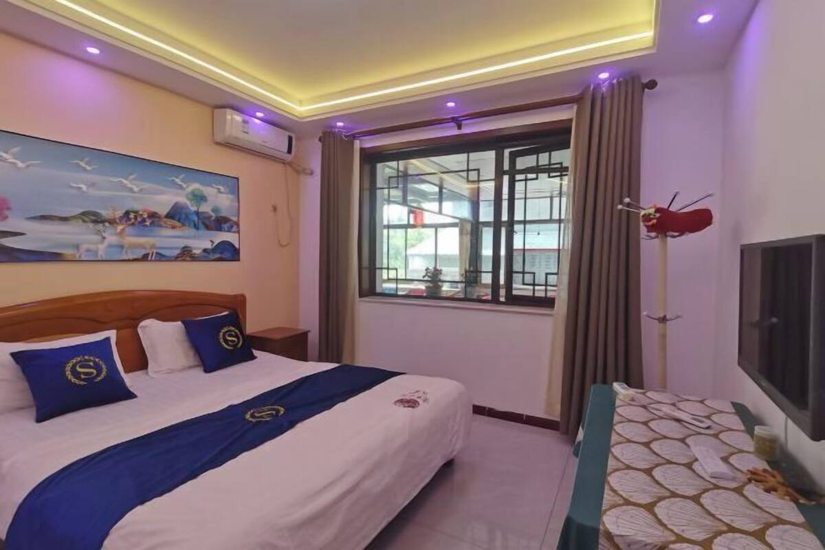 A hotel room with a bed, a painting on the wall, a window with curtains, and a television on the wall.