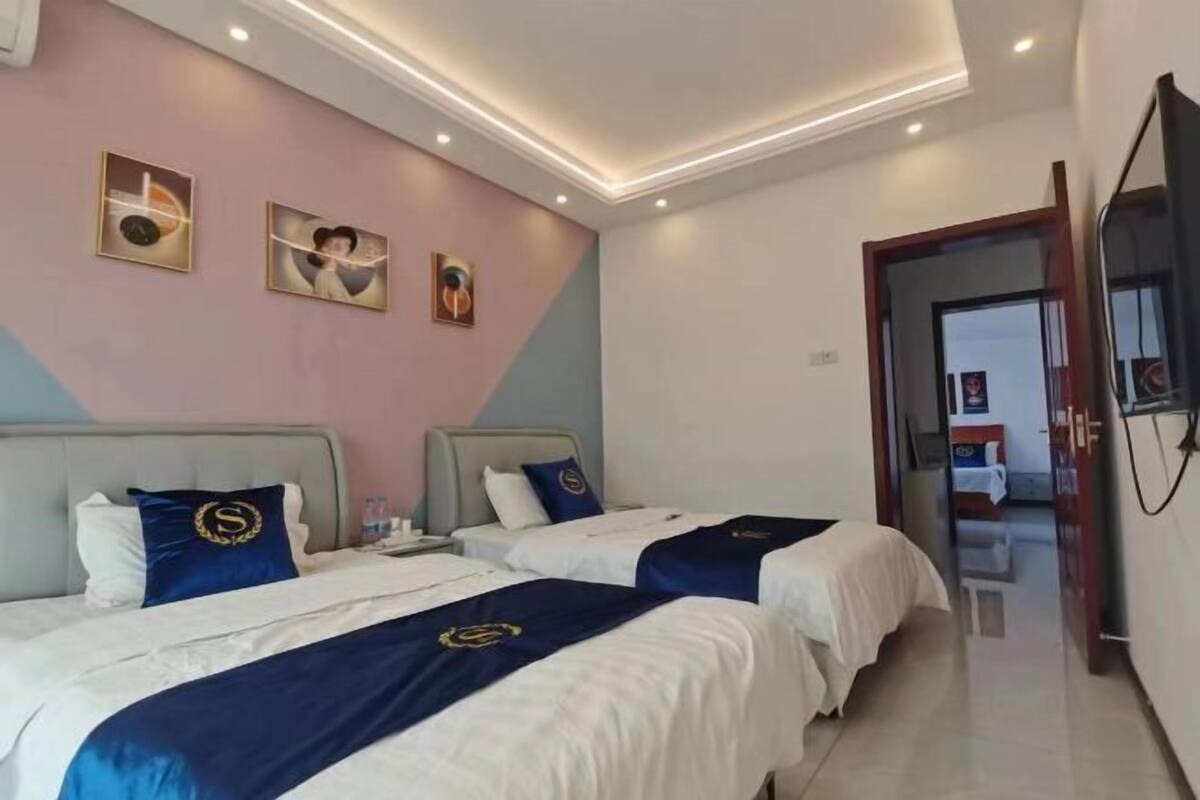A hotel room with two beds, each with a blue bedspread and matching pillows. There are wall-mounted artworks and a television on the wall.