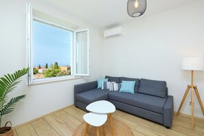 Ett modernt vardagsrum med en grå soffa, ett litet vitt bord, en golvlampa och utsikt över havet genom ett stort fönster.