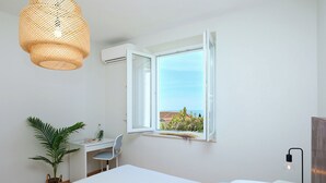 A bedroom with a bed, a desk, a chair, a potted plant, and a window with an open view.