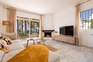 Una sala de estar moderna con sofá, televisión, chimenea y balcón con vista a árboles.