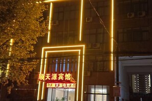 A building with illuminated signage, a tree in front, and a window with blinds.