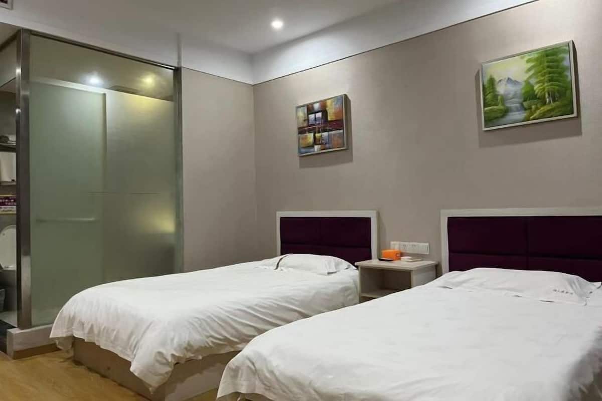 A hotel room with two beds, a glass shower enclosure, a small table with a cup, and artwork on the walls.