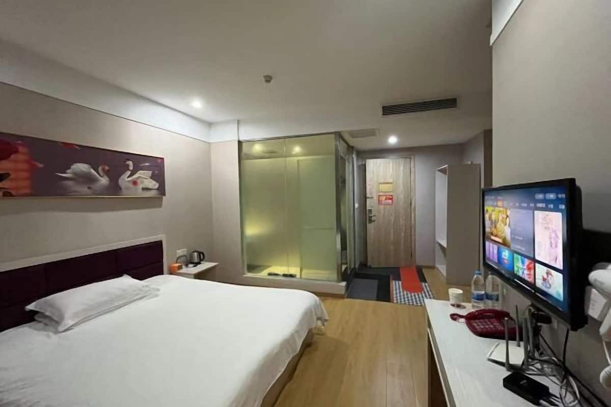 A hotel room with a bed, a desk with a computer, a TV, and a glass-enclosed shower area.