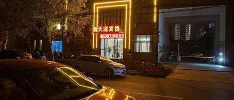 Una calle nocturna con coches estacionados al borde y un edificio con letreros iluminados.
