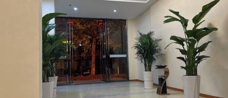 A hotel lobby with a modern design, featuring a glass entrance, potted plants, and a chandelier.