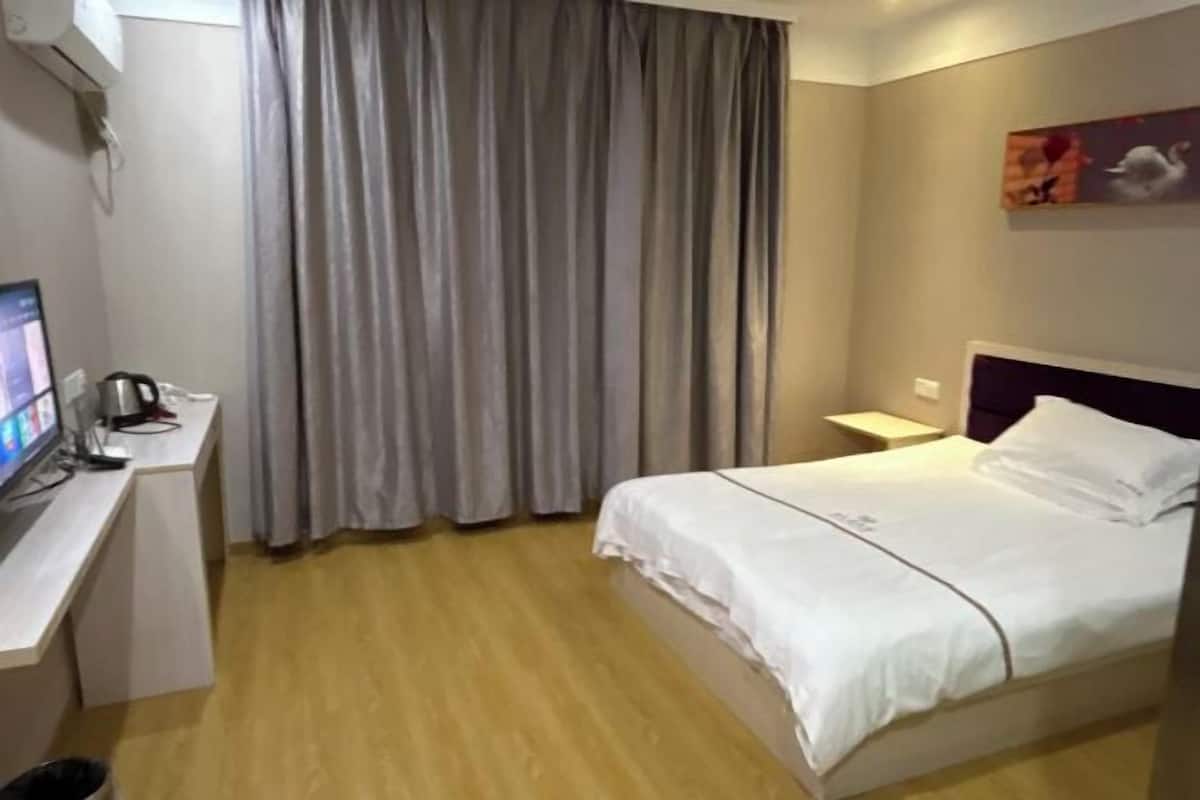 A hotel room with a bed, a desk with a computer, a window with curtains, and a wall-mounted artwork.