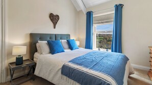 Un dormitorio con una cama grande, una mesita de noche con lámpara, un aparador y vistas al exterior a través de una ventana con cortinas azules.