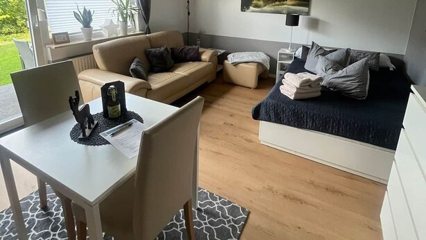 A modern living room with a dining table, two sofas, a ceiling fan, and a large window with a view of greenery.