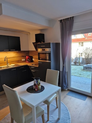 Uma cozinha compacta com eletrodomésticos modernos, área de refeições com mesa e cadeiras, e vista para o exterior por uma janela ampla.