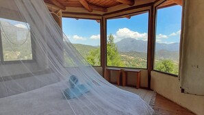 A room with a bed under a wooden ceiling, large windows with a view of mountains, and a mosquito net.