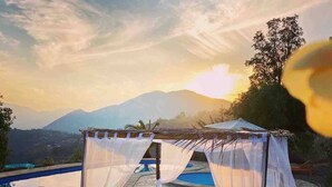 A wooden bench under a white canopy with a pool and mountains in the background.