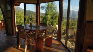 A wooden dining table with wicker chairs in a room with large windows overlooking a mountainous landscape.