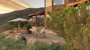 A rustic cabin with a large window, a patio with a table and chairs, and a view of the surrounding hills.
