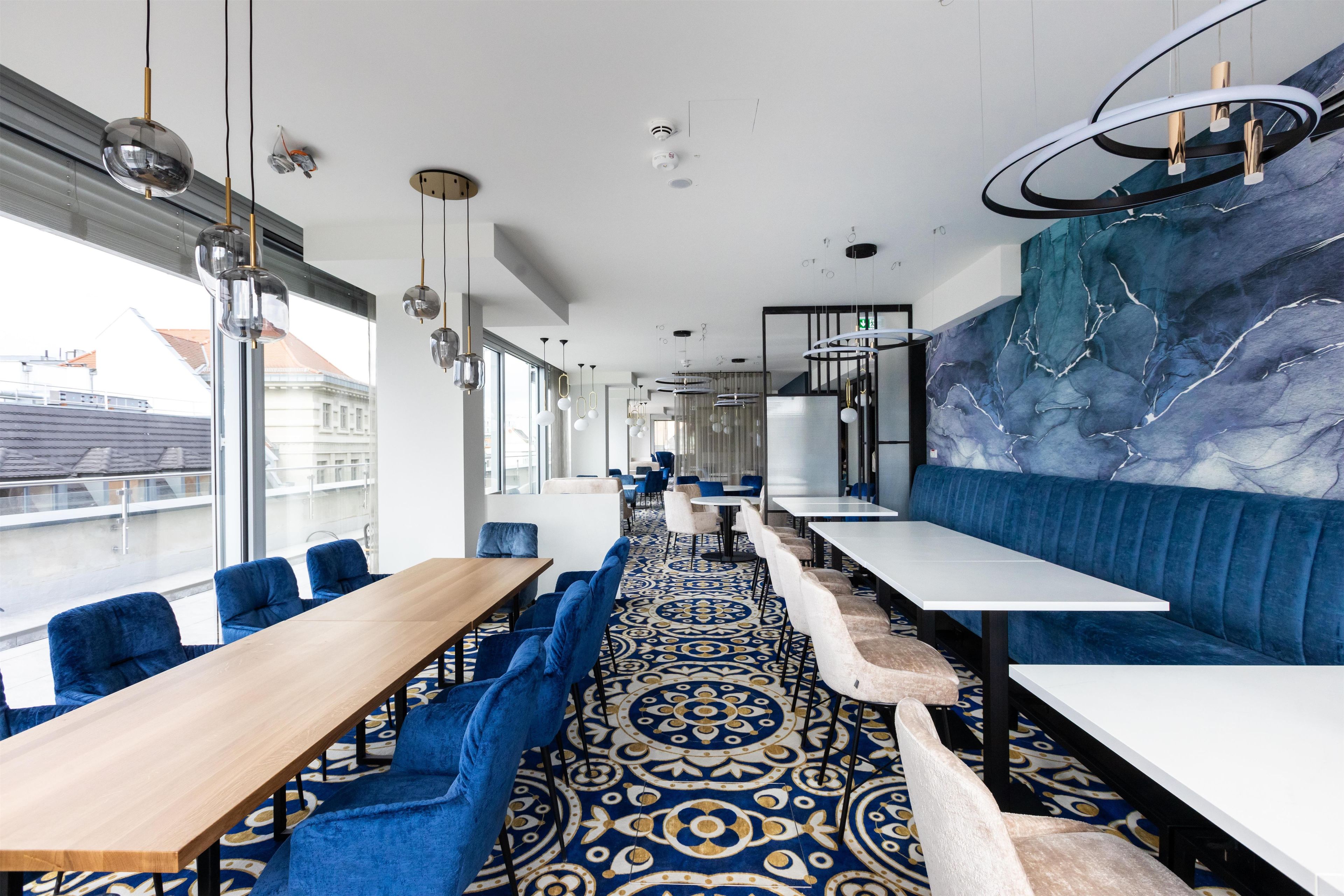 Un comedor moderno con mesas largas y asientos tapizados en azul, una alfombra estampada y un amplio ventanal con vistas a edificios.