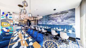 Un restaurante con asientos tapizados en azul, una larga mesa de madera preparada para una comida, y una pared de mármol con un mural.