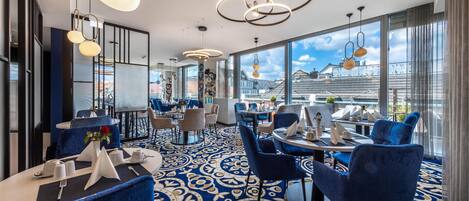 Un restaurante moderno con sillas tapizadas en azul, alfombra estampada y grandes ventanales con vistas a la ciudad.