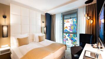 A hotel room with a large bed, a desk with a laptop, a chair, and a window with colorful artwork.