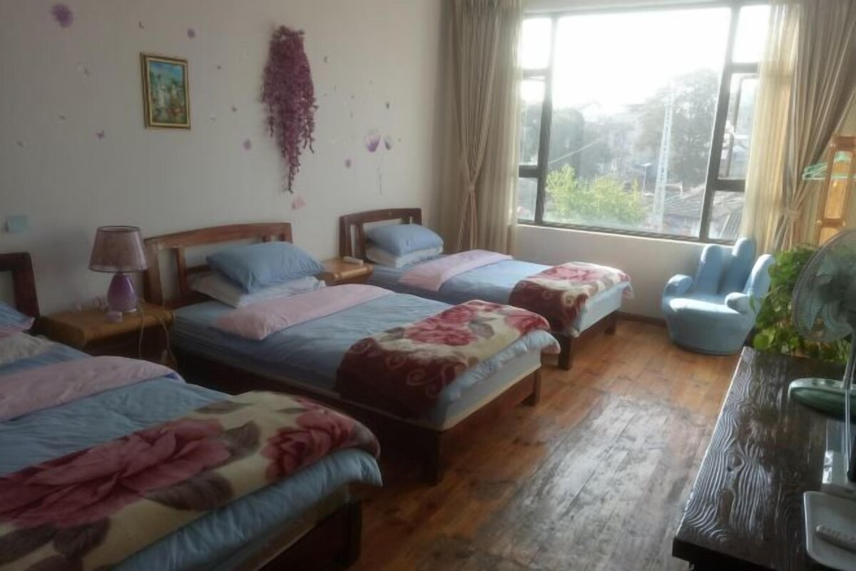 A room with three beds, a wooden floor, a window with curtains, a chair, and a desk with a fan.