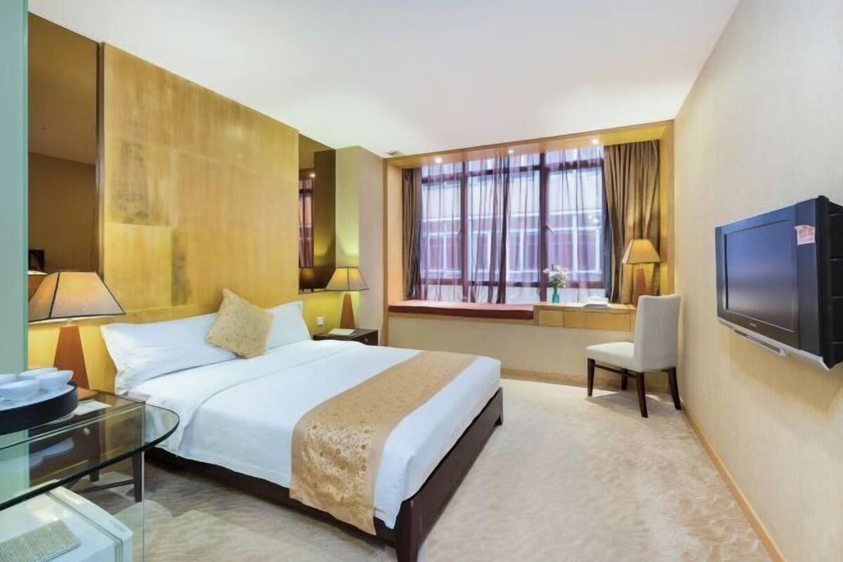 A hotel room with a bed, bedside tables, a TV, a window with curtains, and a desk with a chair.