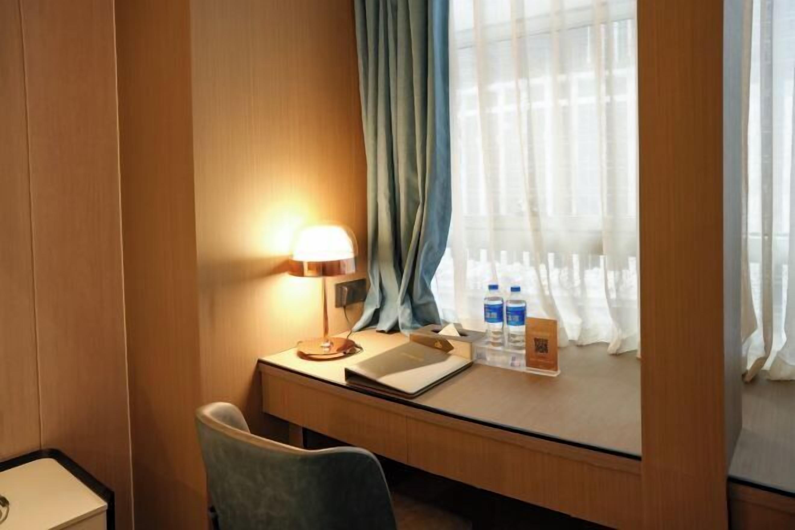 Habitación de hotel con escritorio, lámpara, silla y ventana con cortinas.