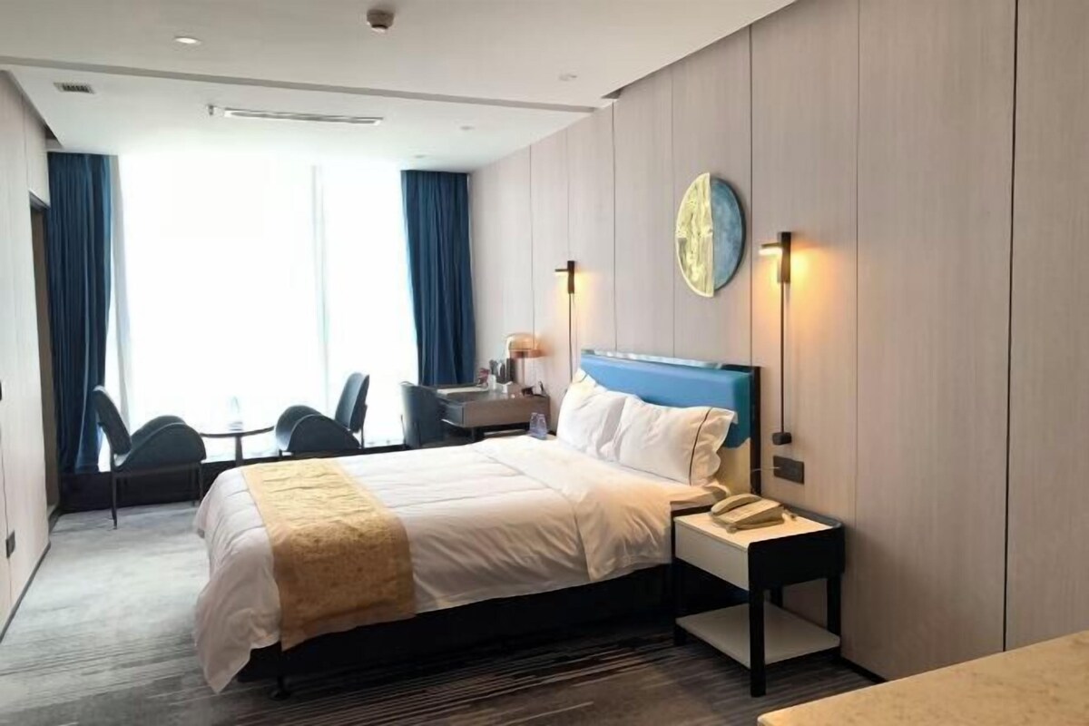 A hotel room with a bed, bedside table, lamp, and a view of the outside.