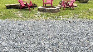 Drie Adirondack stoelen rond een vuurkuil van steen in een gazon, met een grindpad op de voorgrond en een bebost gebied op de achtergrond.