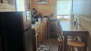 Een kleine keuken met houten kastjes, een koelkast, een eettafel met stoelen en een hanglamp.