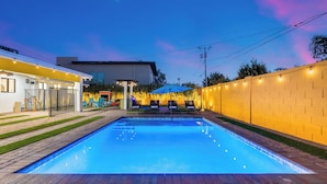 En swimmingpool med murstenskant, omgivet af en gul mur og en hvid bygning med en overdækket terrasse.
