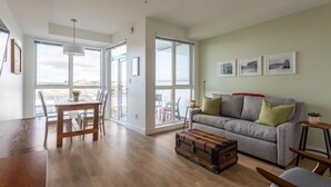 Amplia sala de estar con una mesa de comedor de madera, un sofá gris con cojines verdes, un baúl antiguo y ventanas grandes con vista a la ciudad.