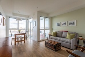 Ein geräumiges Wohnzimmer mit einem Holzespeisetisch, einem grauen Sofa mit grünen Kissen, einem Vintage-Koffer und großen Fenstern mit Blick auf die Stadt.