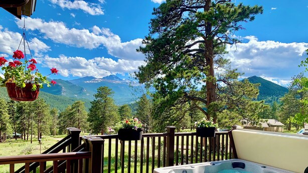 Une terrasse en bois dotée d’un jacuzzi, entourée d’une balustrade en bois, offrant une vue sur un paysage montagneux avec des arbres et un ciel dégagé.
