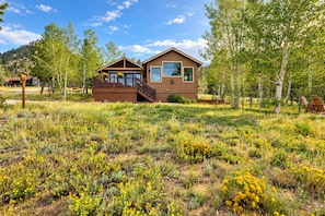 Une cabane à deux étages dotée d’un balcon, entourée d’un paysage naturel où abondent les fleurs sauvages et les arbres.
