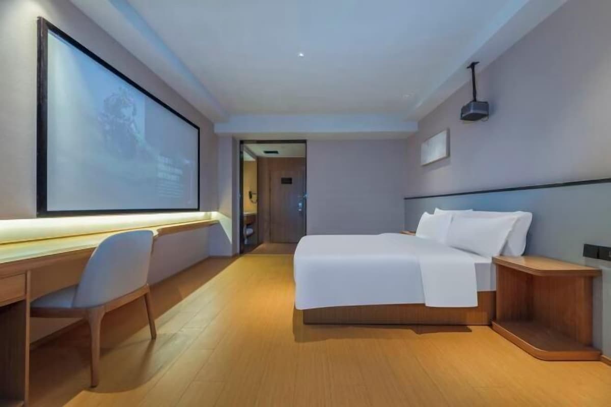 A modern hotel room with a large bed, a desk with a chair, and a large screen on the wall.
