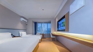 Ein Hotelzimmer mit zwei Betten, einem Schreibtisch, einem Sessel, einem an der Wand montierten Fernseher und einem Fenster mit Vorhängen.