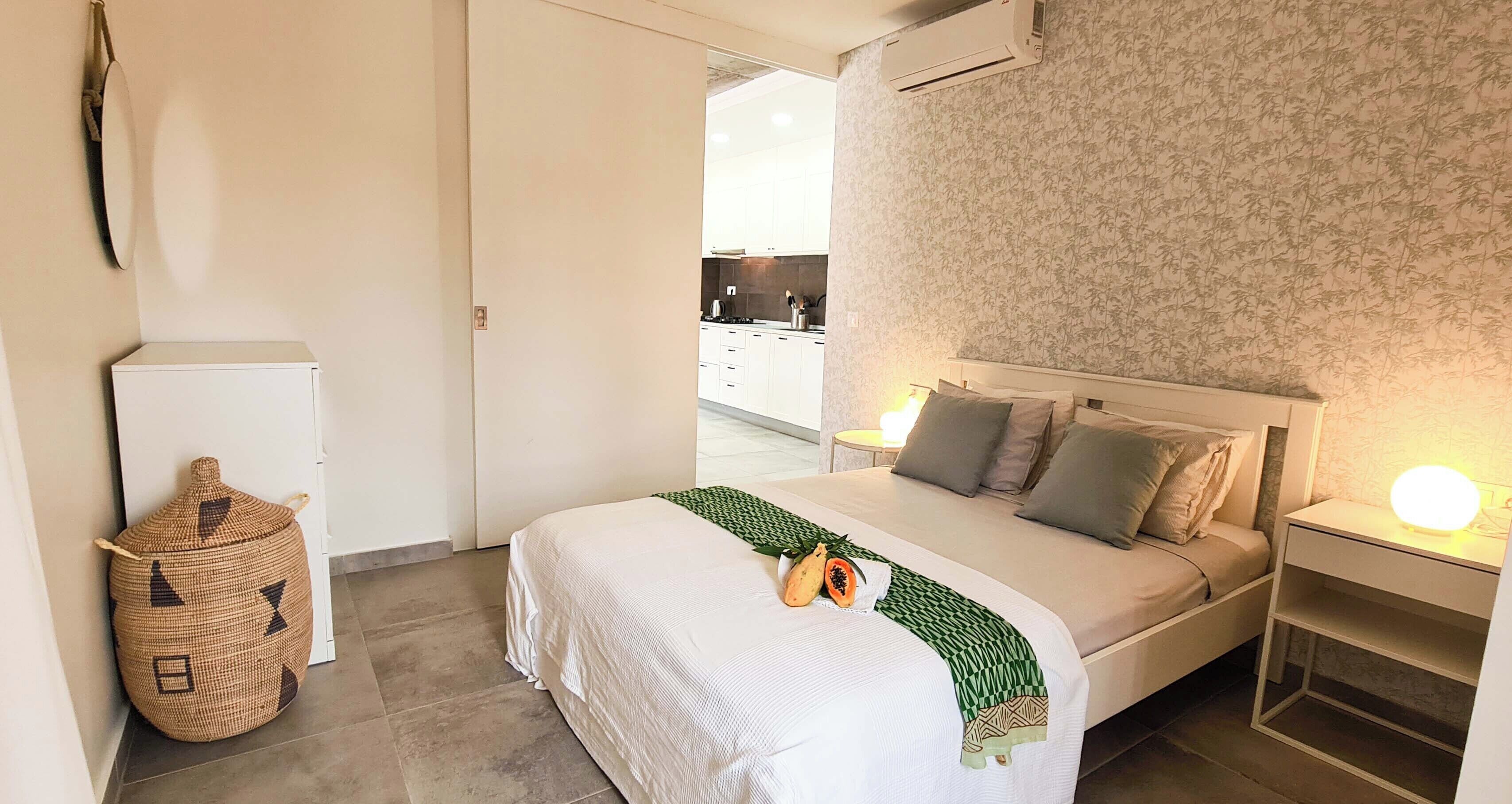 Une chambre à coucher avec un lit, des tables de chevet, un climatiseur fixé au mur et, au fond, une cuisine visible.