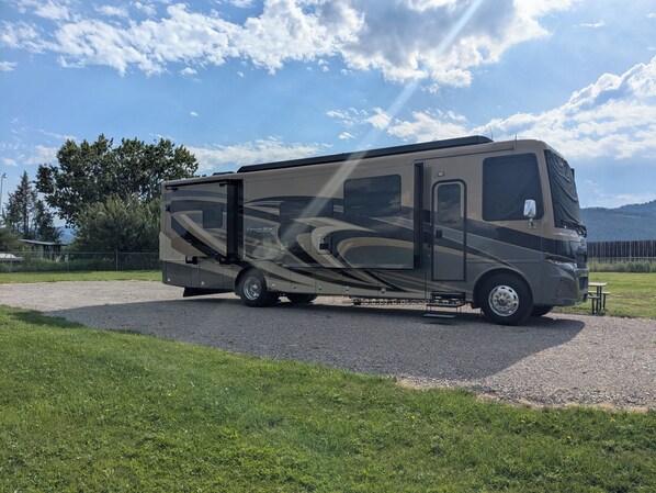 A recreational vehicle parked on a gravel path with a grassy area and trees in the background. - Single R.V. Pad with full hook ups. Located minutes from Flathead Lake! (Kalispell)