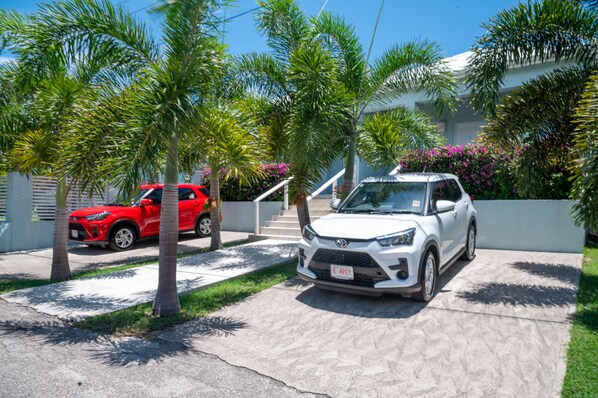 A residential driveway with two cars parked, palm trees, and a house with steps leading to a door. - Trade Winds Cottages #2 (Hodges Bay)