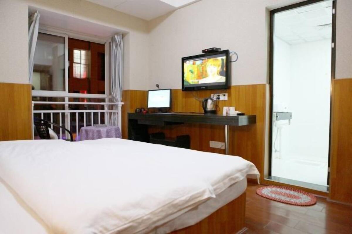 Ein Hotelzimmer mit einem Bett, einem an der Wand befestigten Fernseher, einem Schreibtisch mit einem Computer und einer Glastür zu einem Balkon.