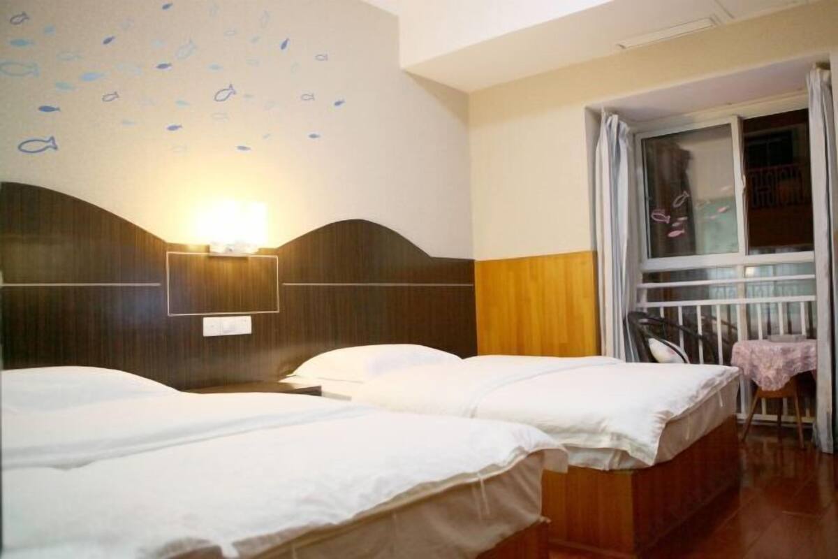 Ein Hotelzimmer mit zwei Betten, einer Wandleuchte, einem Fenster mit Vorhängen und einem kleinen Tisch mit Stuhl.