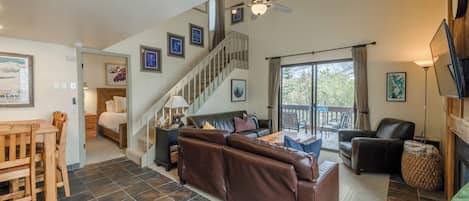 A spacious living area with a brown leather sofa, a dining table with chairs, a staircase, and a large window with curtains.