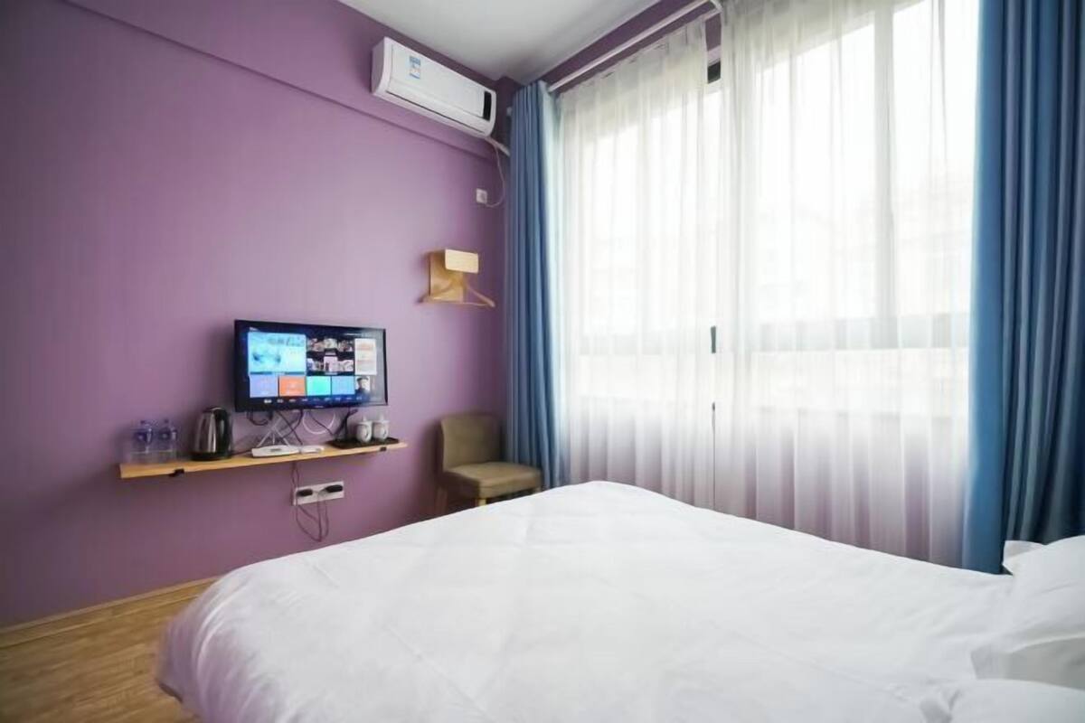 A hotel room with a bed, a television on a shelf, a chair, and a window with curtains.