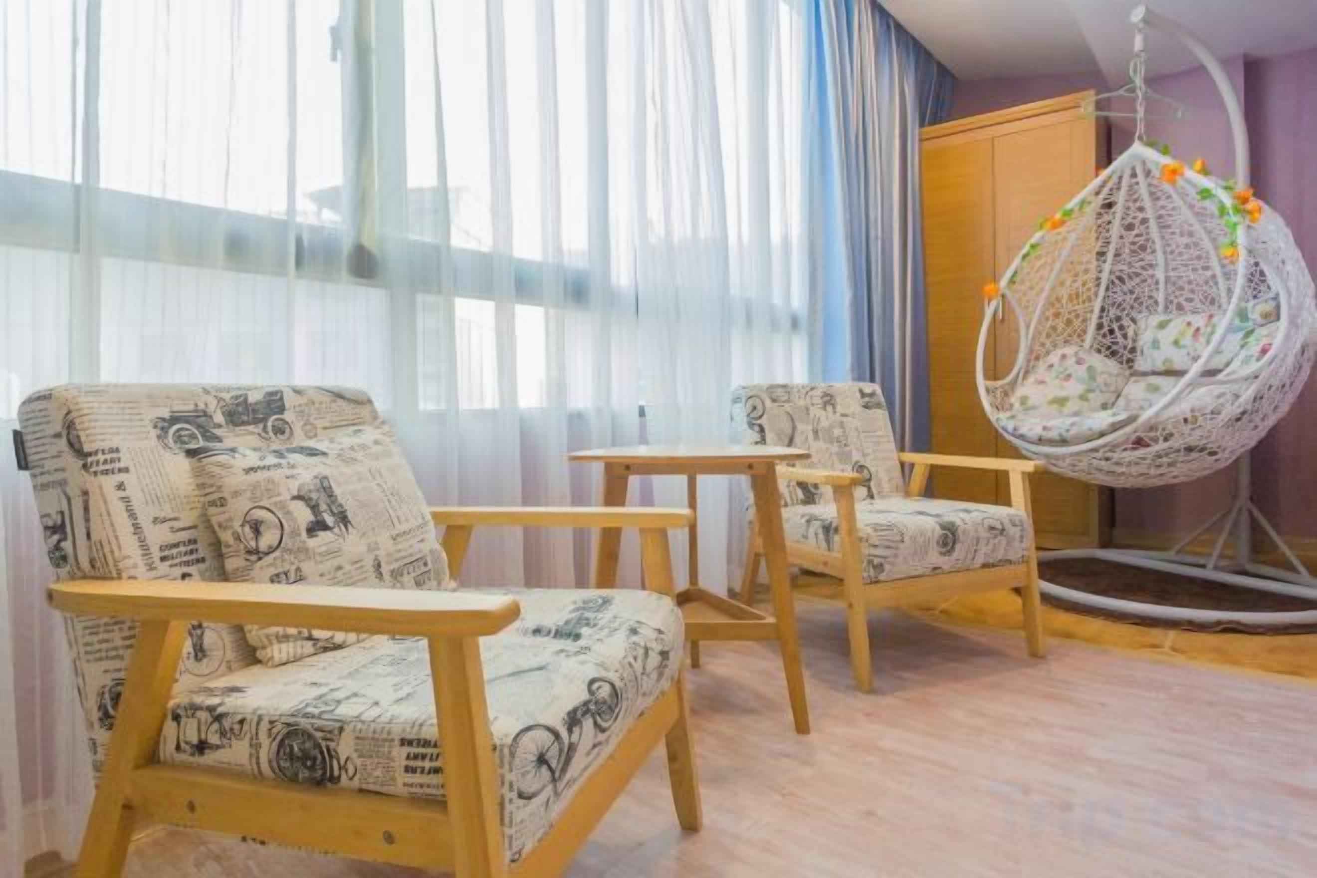A room with two wooden chairs with patterned upholstery, a hanging chair with a floral cushion, and a large window with sheer curtains.