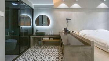 A modern hotel room with a glass-enclosed shower, a round mirror, a bedside table, and a bed with white linens.